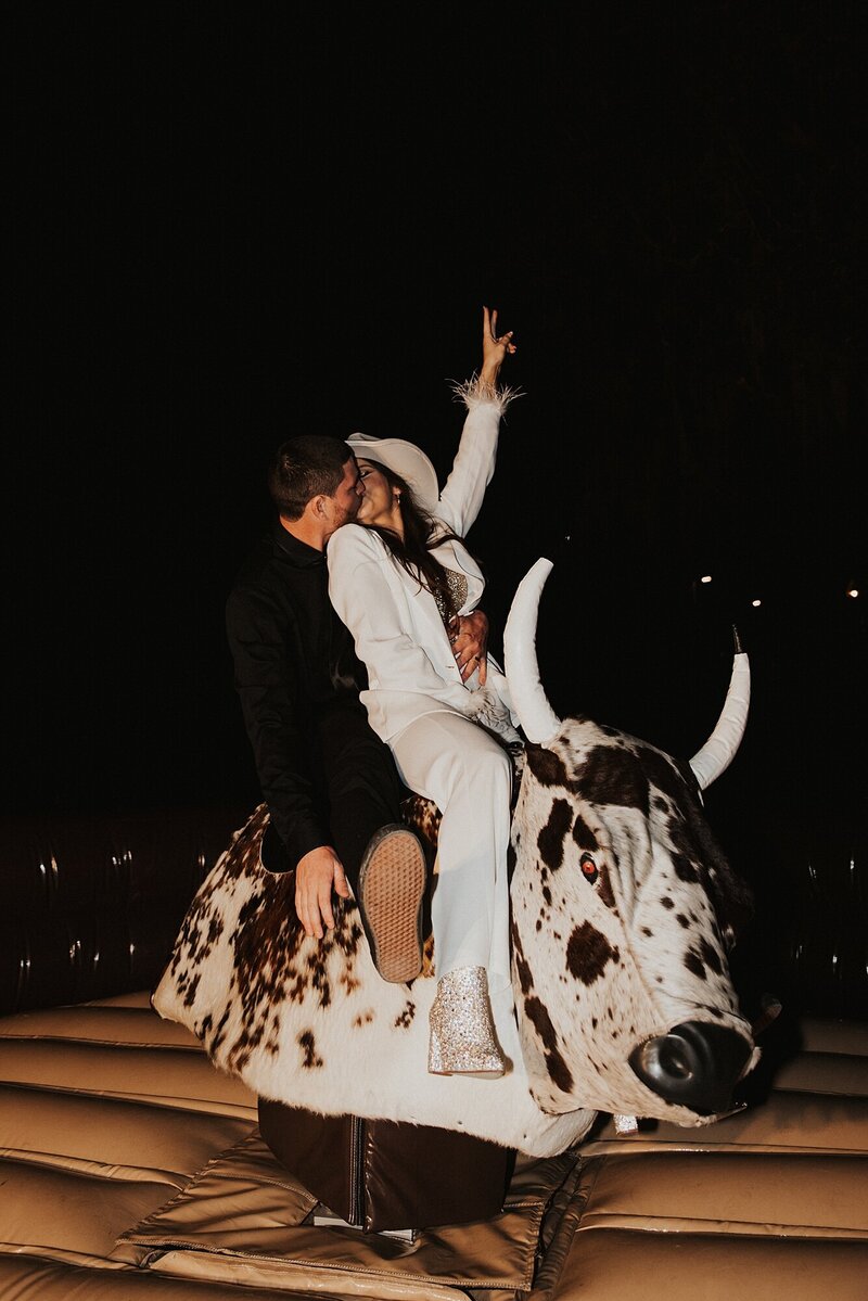 Bride in white jumpsuit and sparkly cowboy hat and boots rides mechanical bull with groom during the Florida elopement