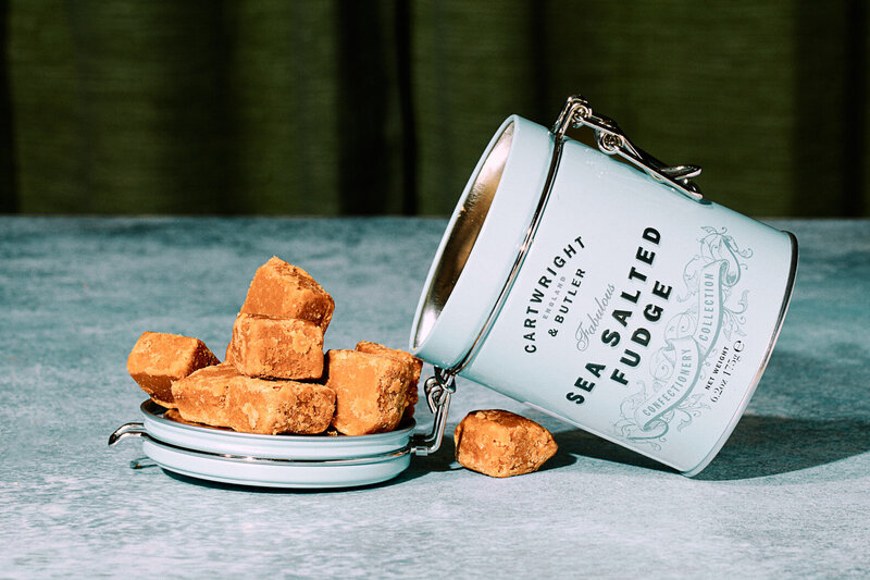 A mouthwatering image of sea salted fudge spilling out of a stylish tin onto a surface. Photographed by Mica McCook, this image captures the rich texture and indulgent appeal of the handcrafted treats.