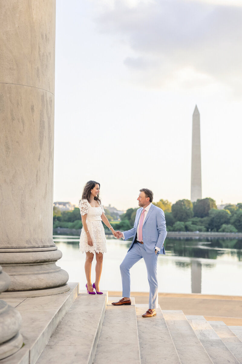 A couple stands hand in hand on stone steps near a tall column, with the Washington Monument reflected in the water behind them. The woman wears a white dress, and the man is in a blue suit with a pink tie. They seem happy and relaxed.