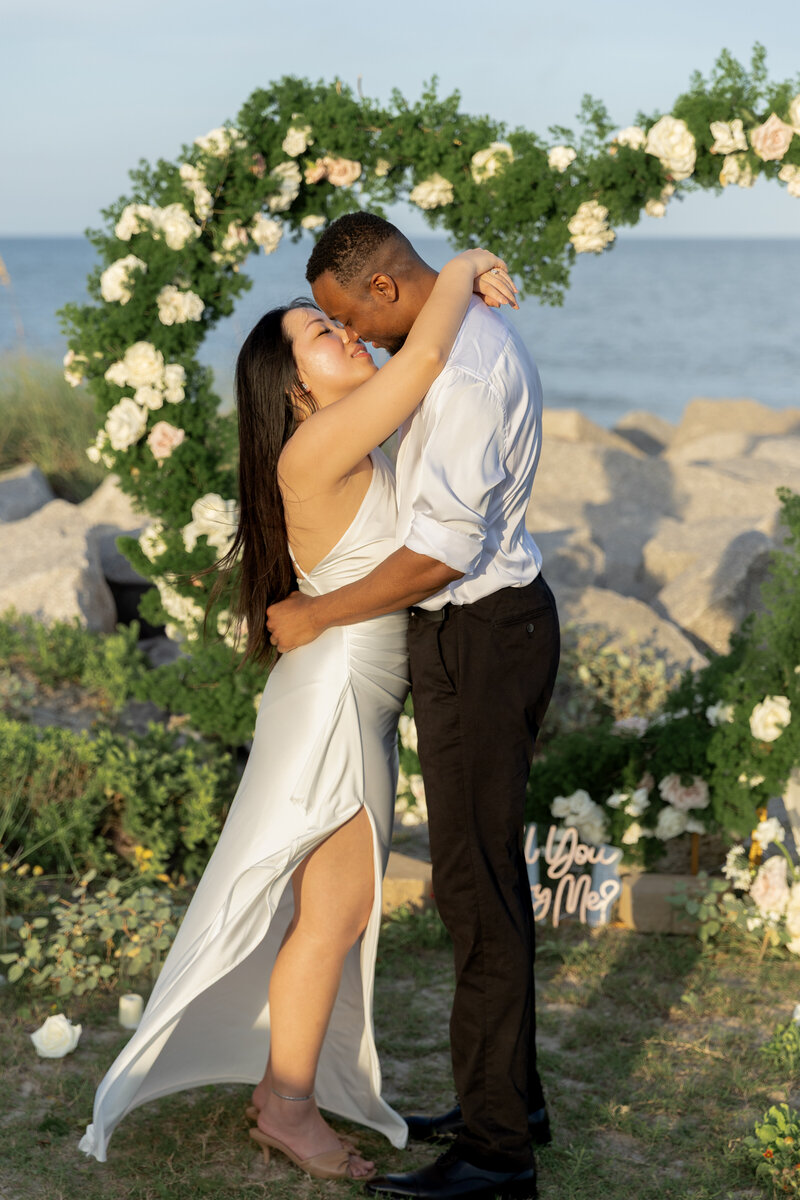 Bride and groom sharing a kiss under a floral arch during a romantic North Carolina beach proposal, photographed by Moments by Hiba.