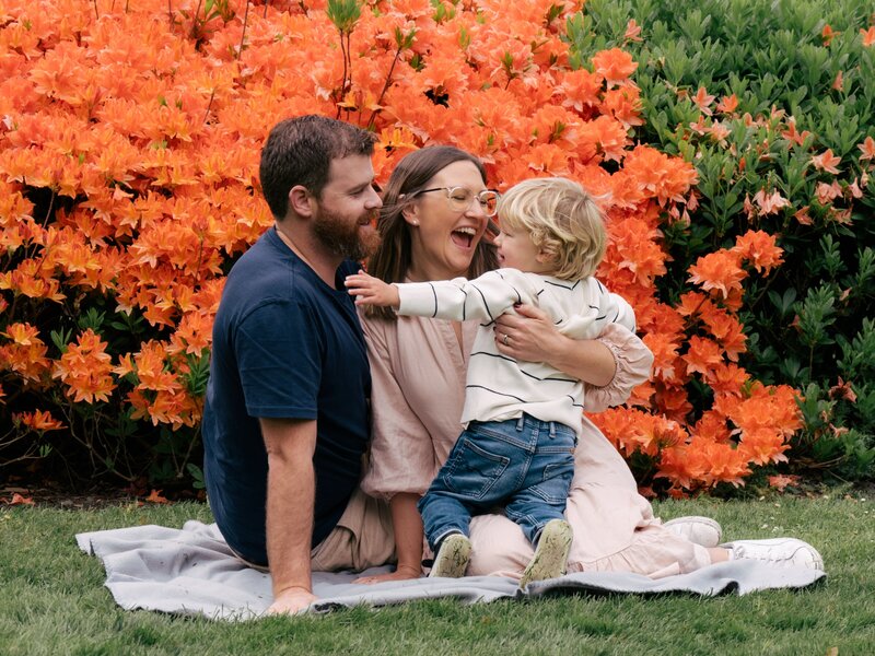 fun family photo of a toddler running into his parents arms. they are on a blanket in front of orange flowers in Christchurch Botanic Gardens.