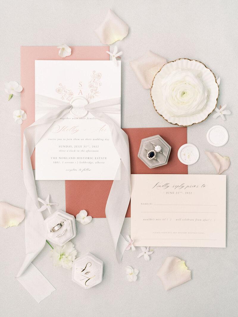 A wedding invitation suite with white, pink, and gray elements, including an invite, RSVP card, envelopes, rings, and flowers arranged on a flat surface by a full-service wedding planner for your perfect day.