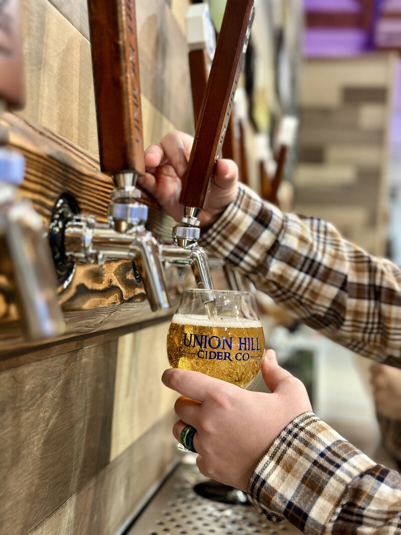 A man pouring hard cider from the tap into a glass that says Union Hill Cider Co. The glass is almost full of yellow-orange cider with some bubbles on top.