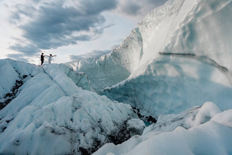 A bride and groom in their formal wedding attire stand atop a large sweeping ice cliff as they trek to their ceremony location during a two day luxury elopement on a glacier in Alaska.  They are high on the icy hill, and look tiny silhouetted agains the sky and setting sun. 
