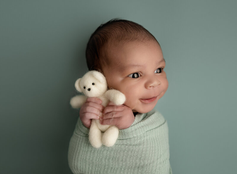 Baby boy in green wrap, holding a felted bear and smiling. 