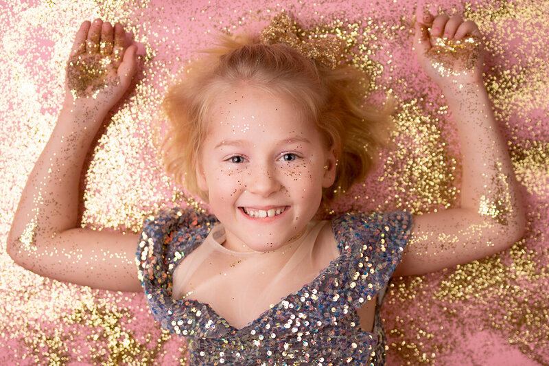 Joyful portrait of a young girl lying on a pink backdrop, surrounded by shimmering gold glitter and wearing a sparkly dress. Captured in Overland Park, Kansas, this vibrant and playful setup is perfect for families seeking creative and magical child photography in the Kansas City metro area