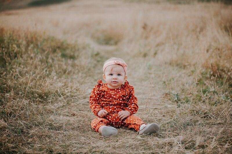 A baby girl sits on the brown grass looking at the camera while wearing a polkadot long sleeve red romper and a pink headband with a bow