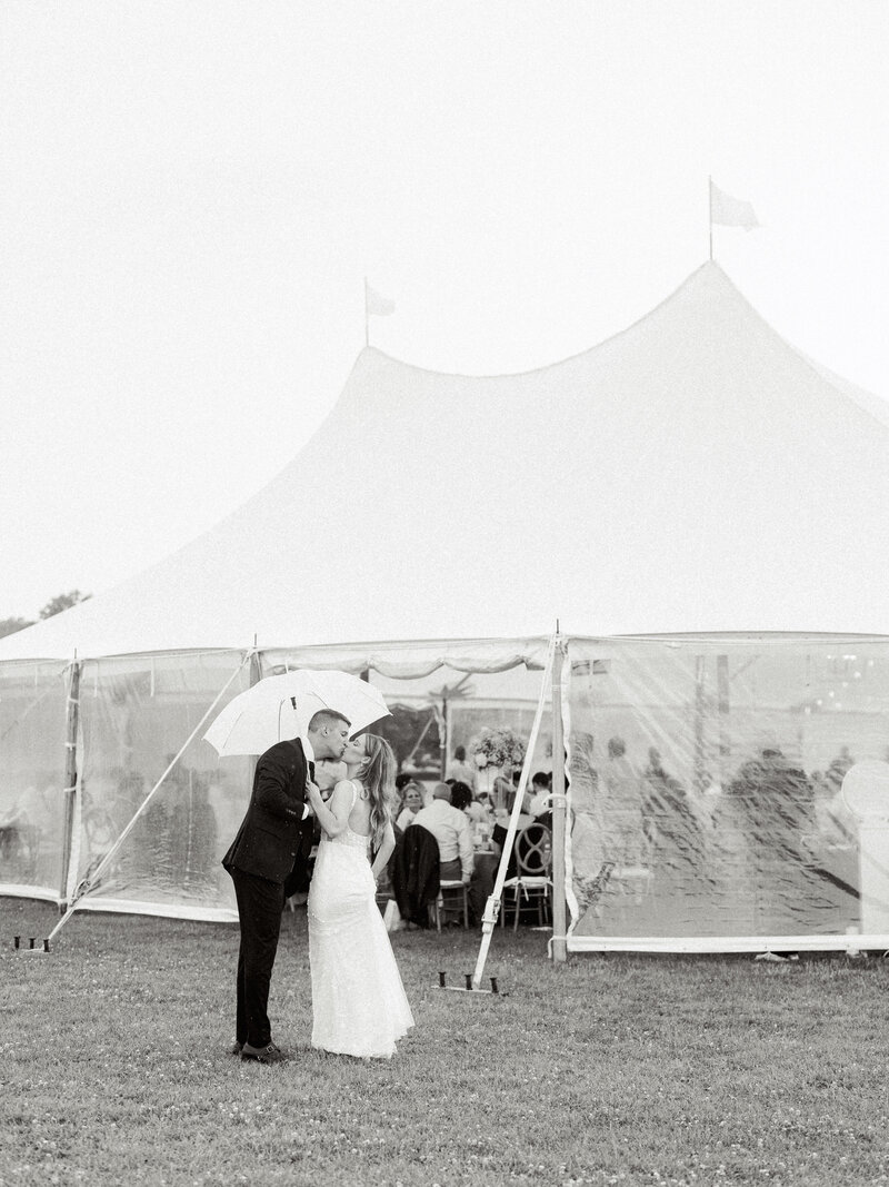 Black and white photo of groom and bride kissing under an umbrella as they're about to enter their tended wedding reception