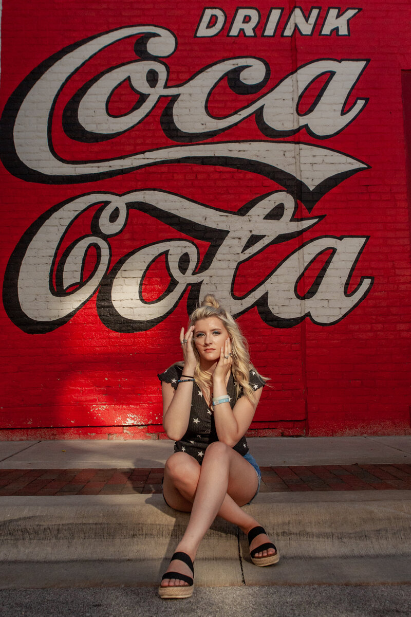 Photo of senior girl sitting on a curb in front of Cola mural where light falls right on the senior.