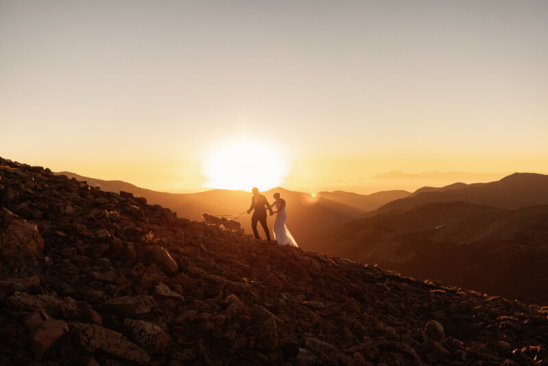 Shannon Lee, a Colorado elopement photographer capturing a couple walking hand in hand against a breathtaking sunset backdrop on a rocky mountain slope.