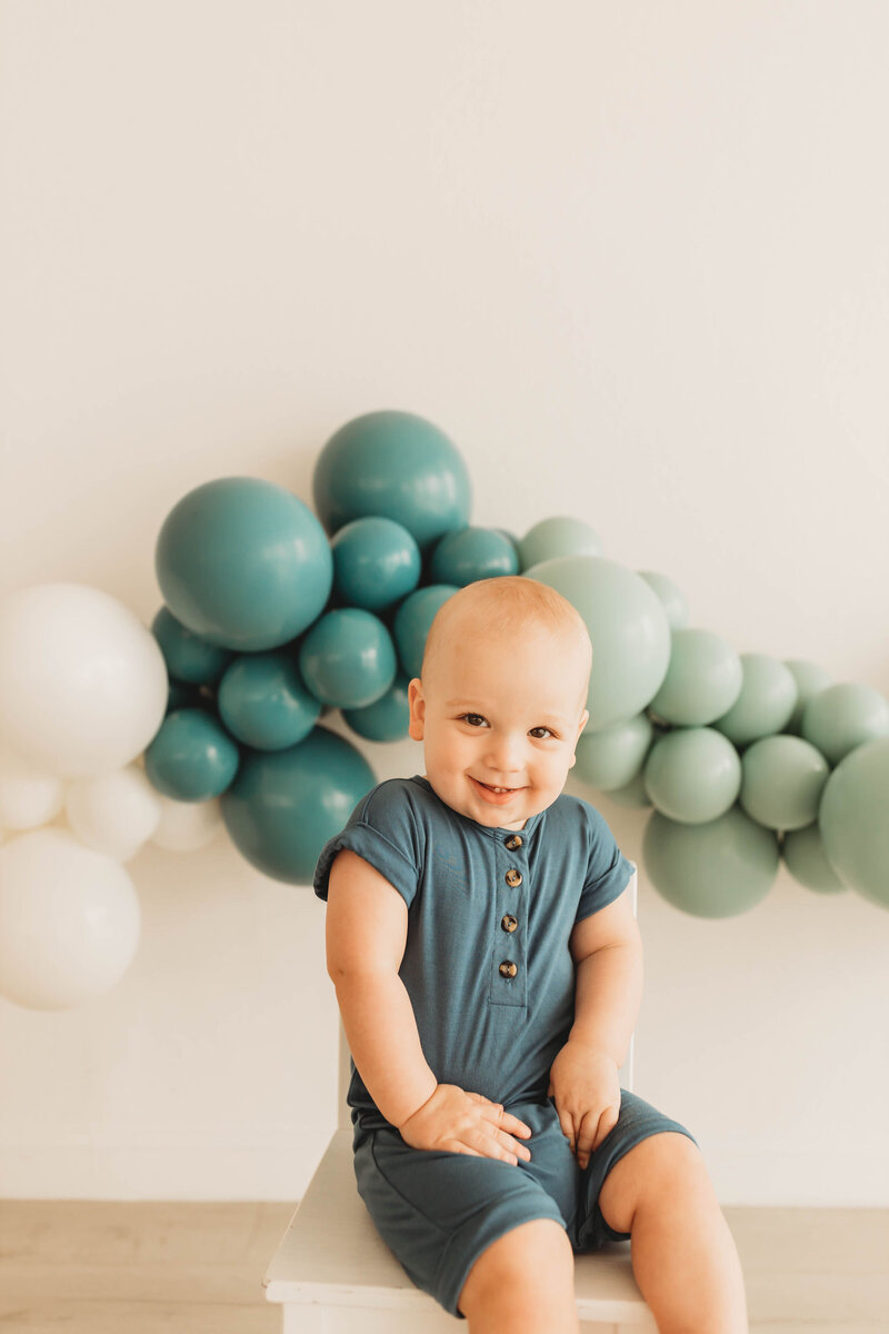 blonde baby boy in a blue onsie in front of balloons in different shades of teal
