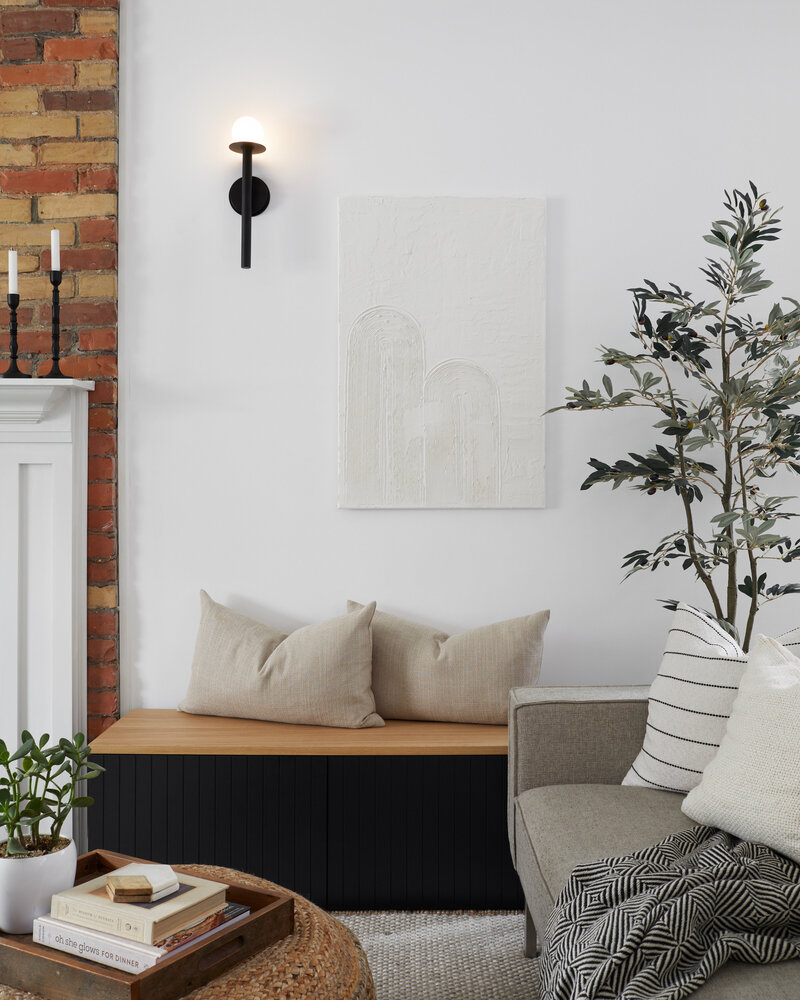 Two off-white decorative pillows on top of a wood bench with black paneling in a bright living room below a white wall-mounted painting and surrounded by potted plants