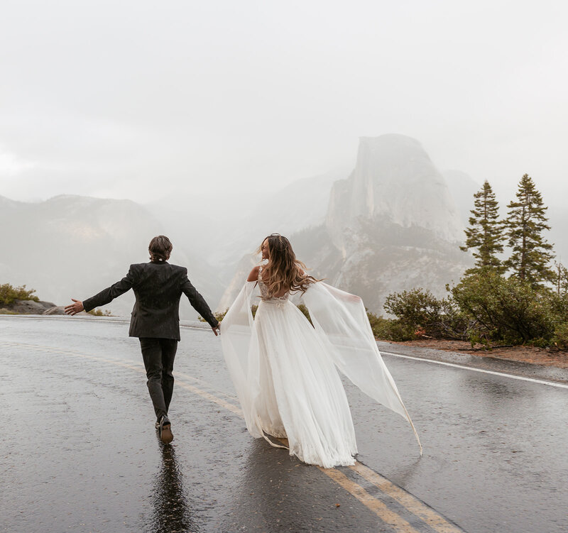 A couple walks hand in hand down a misty mountain road in Yosemite National Park, the bride’s flowy gown billowing behind her as Half Dome peaks through the fog, captured beautifully by Sydney Breann Photography.