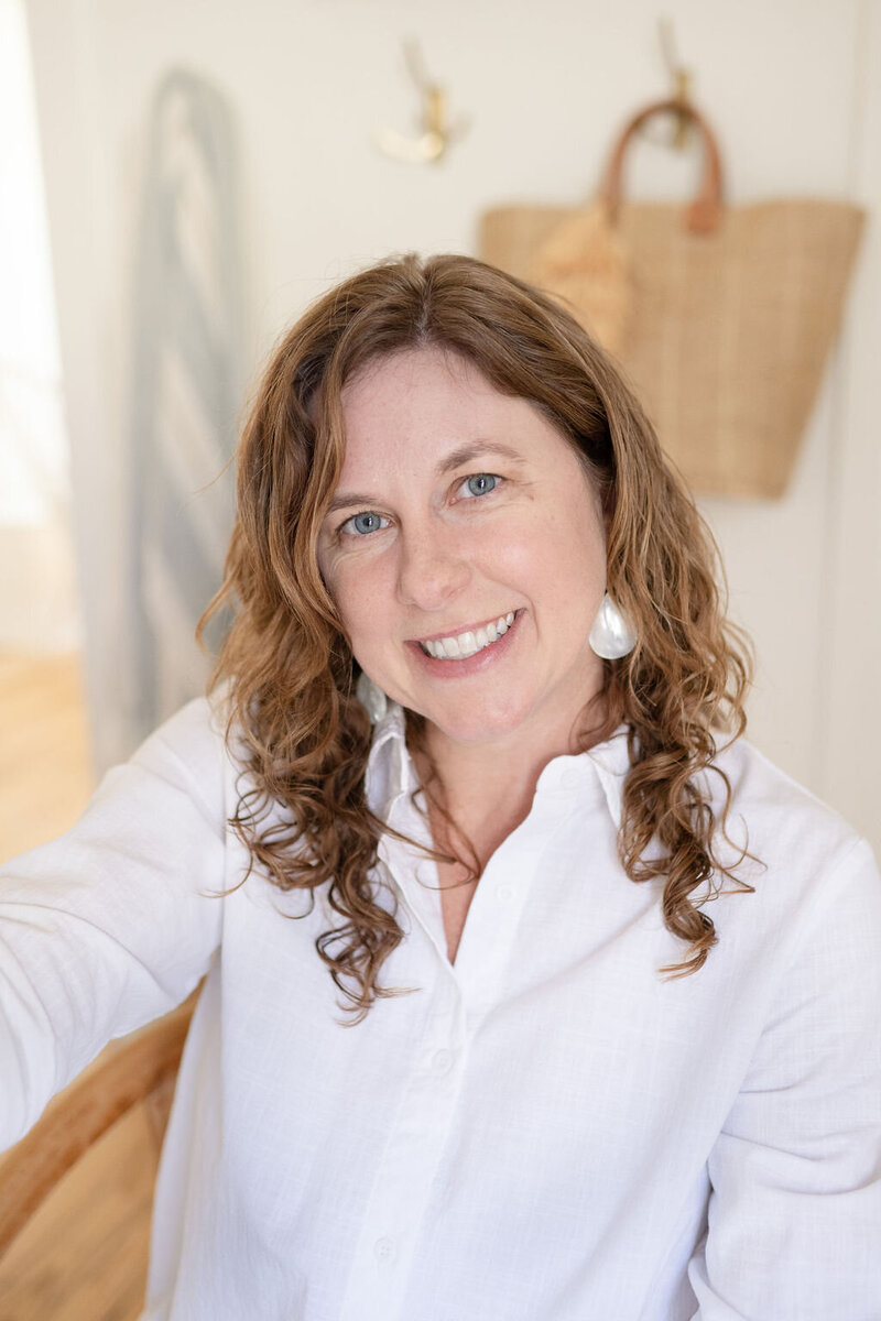 a closeup portrait of Laura, a watercolor artist wearing a white button up and white earrings with a canvas tote bag hanging  on a hook in the background