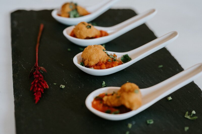 Four grbic tasting spoons with fried appetizers on red-orange sauce, garnished with herbs