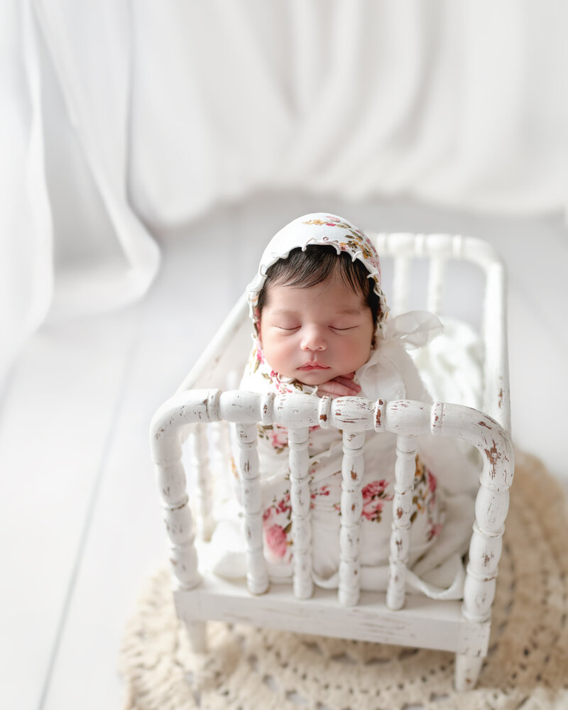Light, airy vintage crib portrait featuring a newborn girl in a floral bonnet and wrap, bathed in soft white tones.