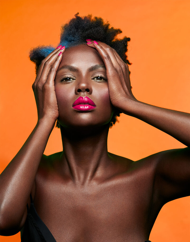 Striking beauty portrait of Lilian against an orange background, showcasing bold makeup and expressive pose, captured by Ylva Erevall.