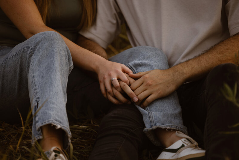 Close-up of a couple sitting in grass, gently holding hands with an engagement ring visible on her finger, both wearing denim.