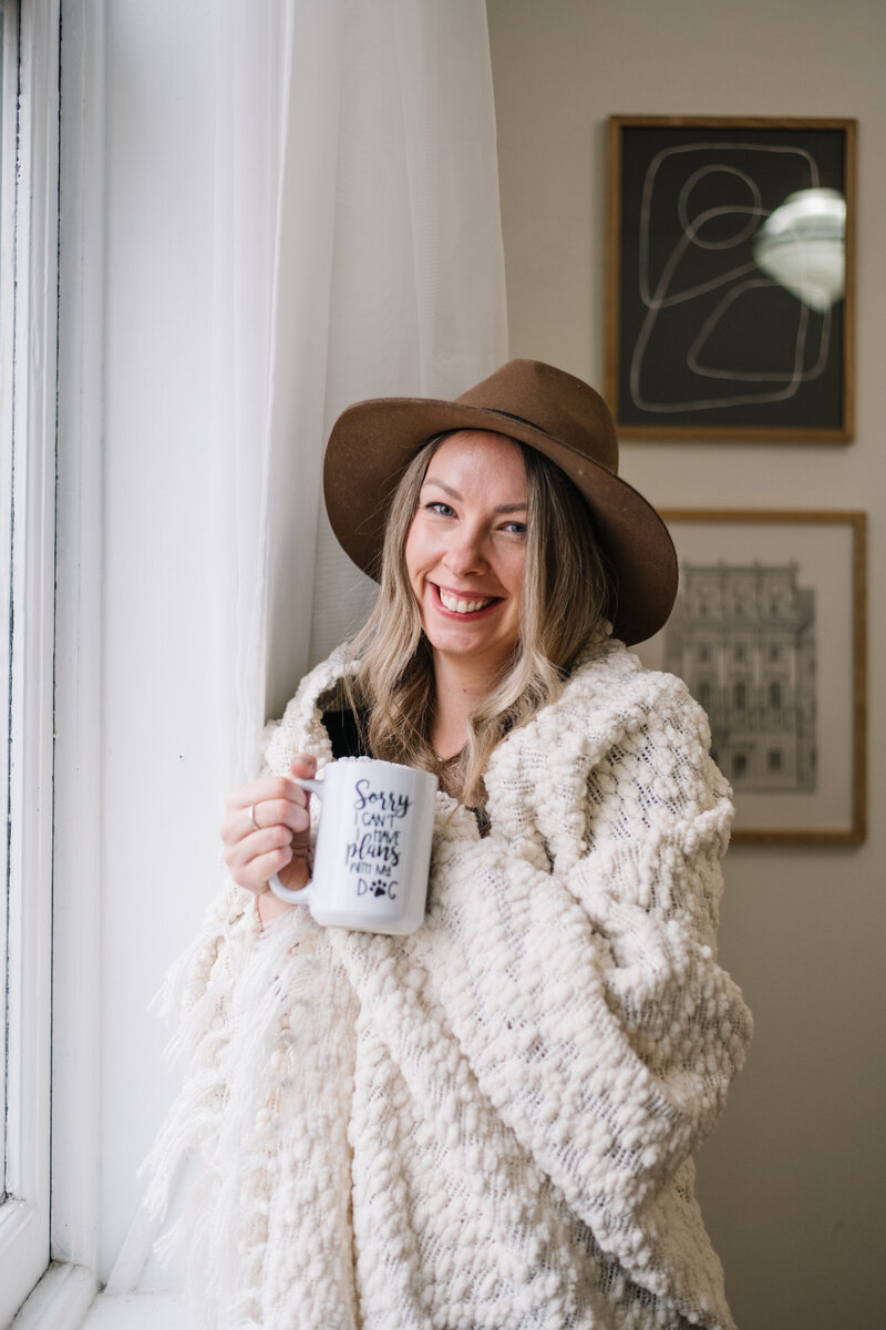 Sammy in a brown hat and wrapped in a white blanket holding a mug