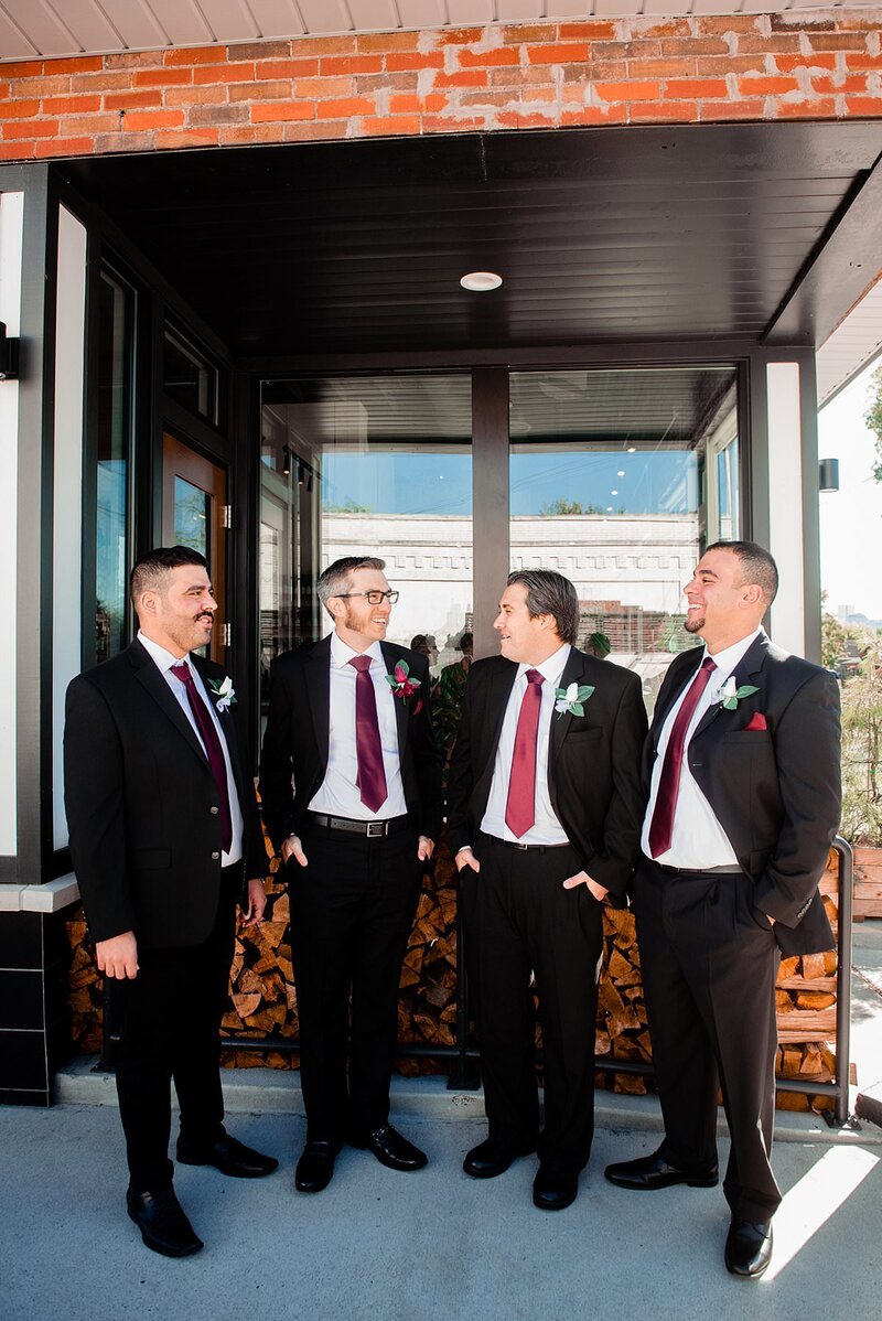 Groom and groomsmen together in front of a brick wall with wood, they are in black suits with red ties