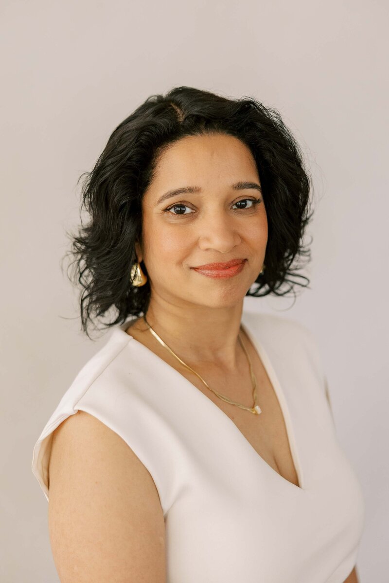 Beautiful Indian woman wearing gold jewelry and an all white outfit and smiling at the camera with a soft smile
