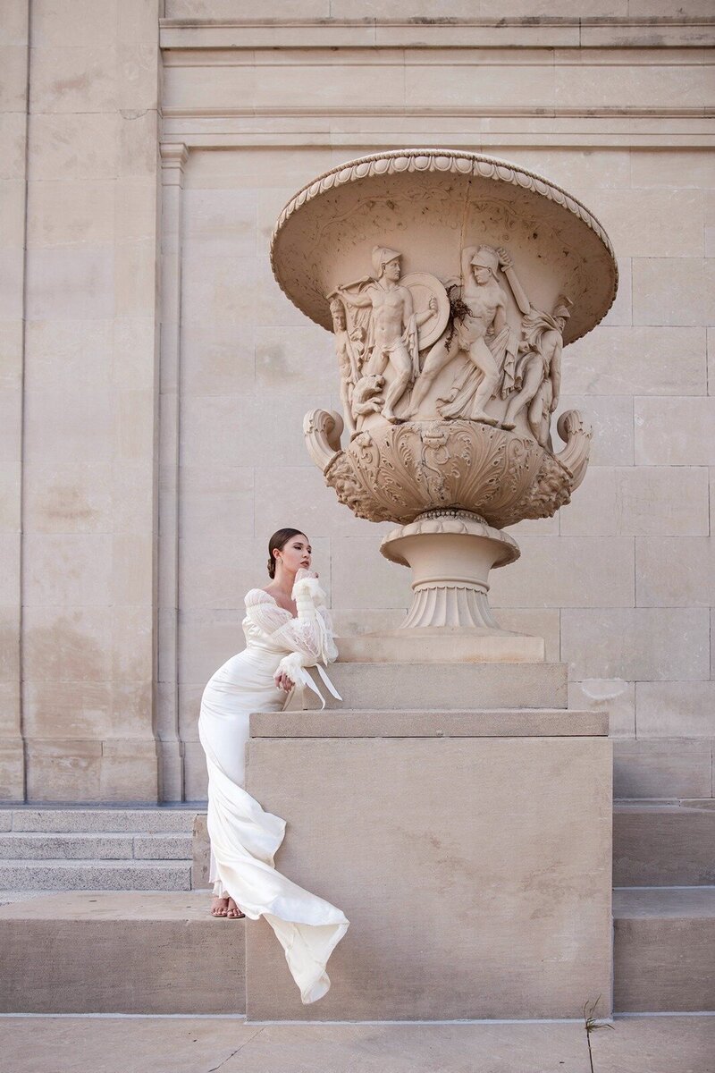 Bridal editorial with wind sweeping dress's train and satin sleeve buttons windswept as she stands by the NOLA vase and exterior architectural facad.