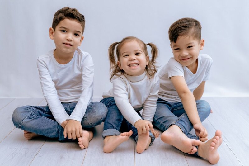  A group portrait of three siblings sitting side-by-side on a light-colored wooden floor against a white background. The older brother on the left has a neutral expression, while the younger brother on the right is smiling. The little girl in the middle, with her hair in two pigtails, is laughing with her mouth open. All three are wearing white long-sleeved shirts and blue jeans.