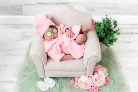 Newborn baby girl in a pink robe with cucumber slices on her eyes, posed on a miniature chair prop with flowers and greenery — spa-themed newborn photography by Misty Dawn Photography in Birmingham, AL.
