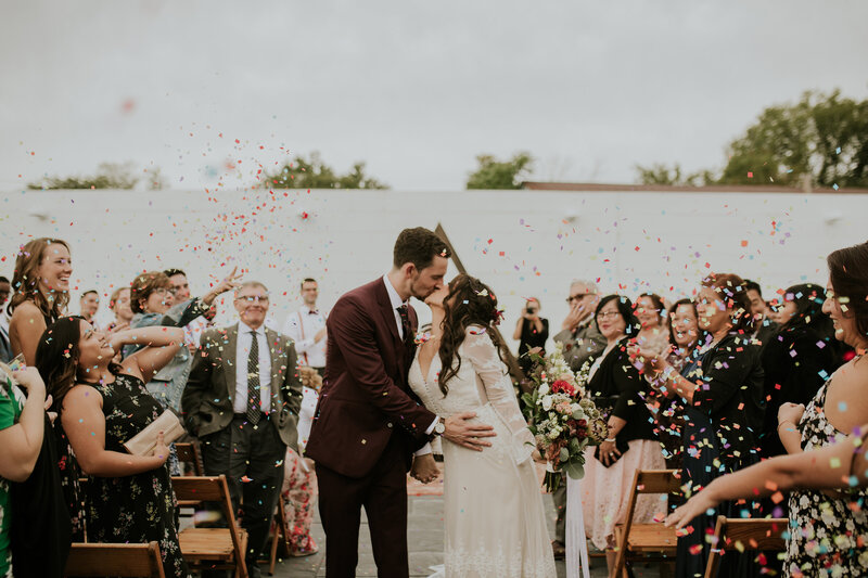 A bride and groom kiss as the guests applaud and throw confeti at their autumn wedding