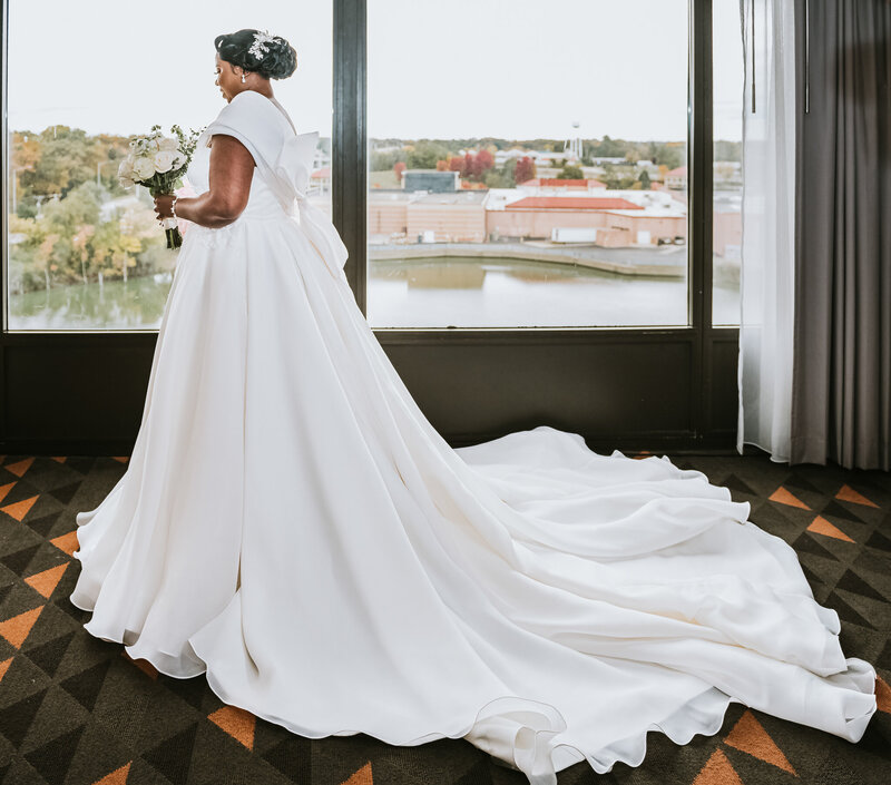 A bride in a stunning white wedding dress with a dramatic long train stands by a window overlooking a cityscape and river. She is holding a bouquet of white roses, her hair styled elegantly in a bun adorned with white flowers. The room has a modern look with large windows and patterned carpet.