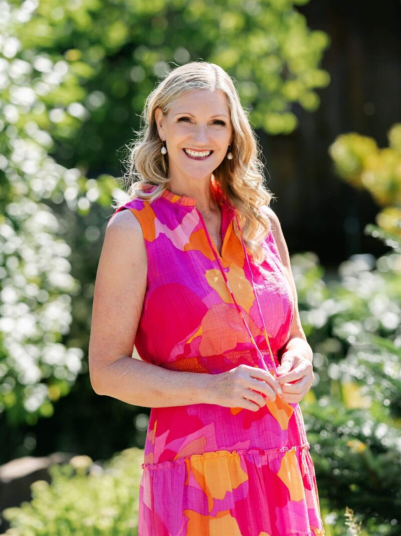 Phillipa Leseberg, an author with long blonde hair, stands outdoors in a bright pink and orange dress, surrounded by vibrant yellow flowers.