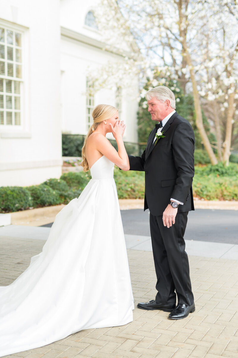 Bride in elegant white gown wipes tears during emotional first look with her father in front of a white venue with blooming trees.