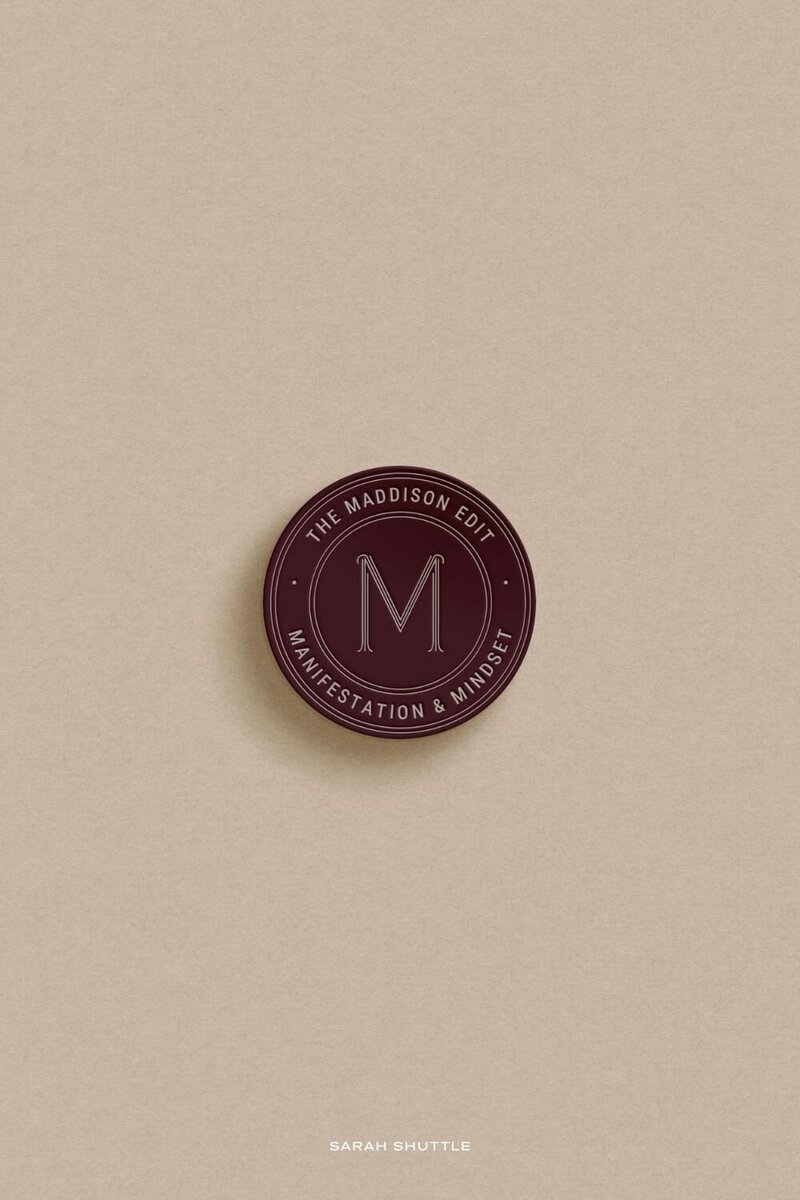Circular burgundy submark logo for The Maddison Edit displayed on a neutral background, adding warmth and sophistication to the brand.