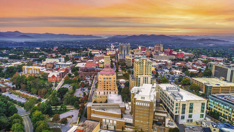 This image shows an overhead view of downtown Asheville, North Carolina, with a pink and yellow sunset about the cityscape in the foreground and mountains in the background.