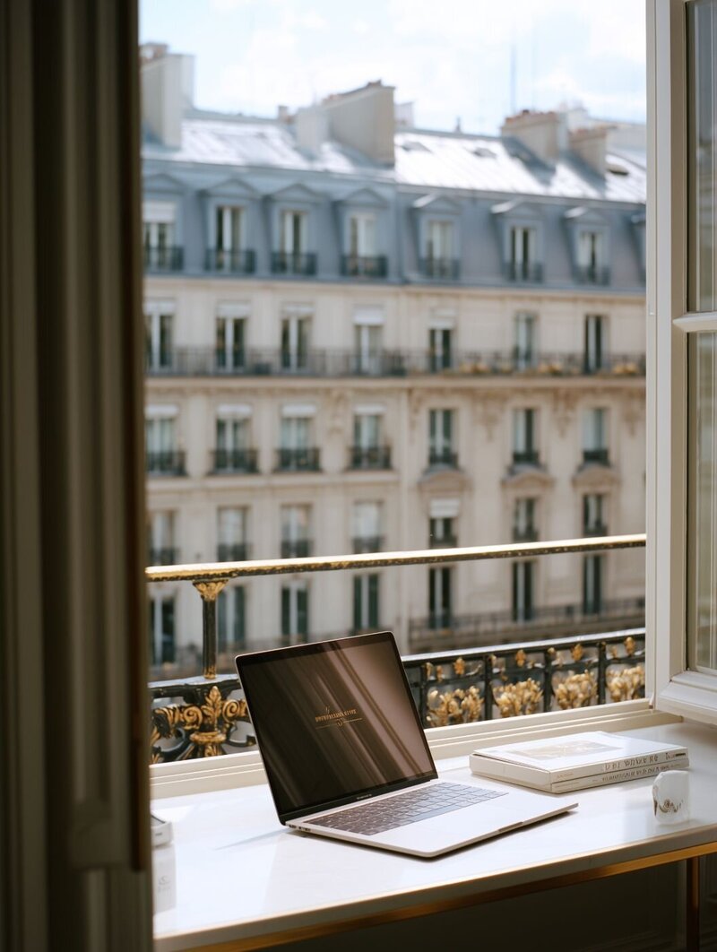 Open laptop on a Parisian balcony windowsill with books, coffee cup and classic Haussmann-style buildings in the background