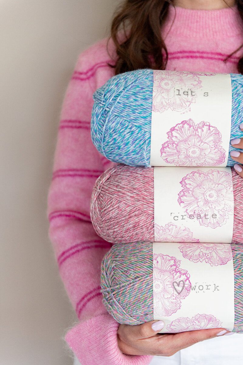 Vrouw met 3 grote bollen wol waar op de labels staat 'let's create heart work'. 