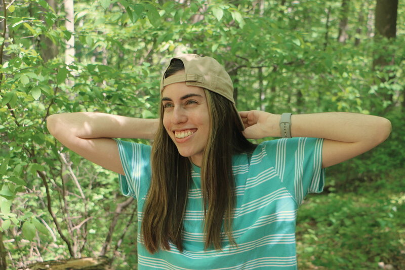 Julia smiling in the woods and looking off camera with her hands behind her head holding the brim of her backwards hat