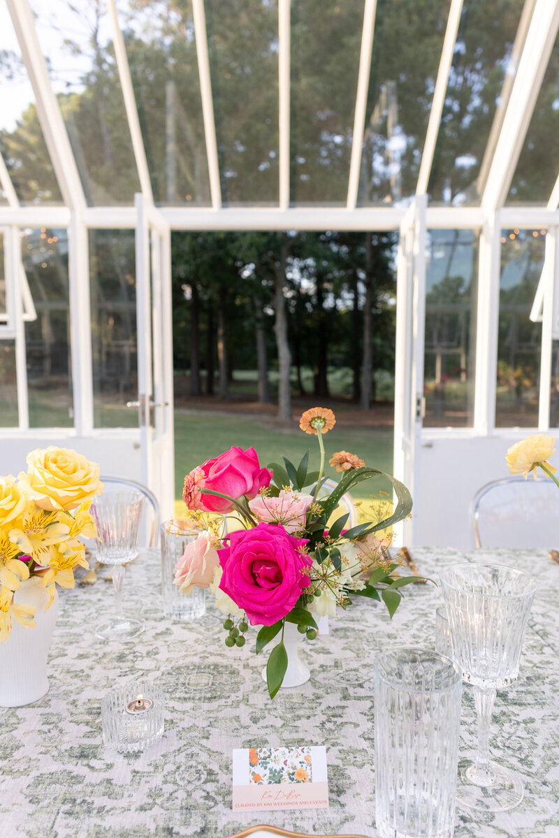 Grey Cove Designs wedding reception table styled with vintage green toile linens, crystal glassware, and a vibrant floral centerpiece featuring pink garden roses and yellow blooms, inside a glass greenhouse with open doors to a lush outdoor view.