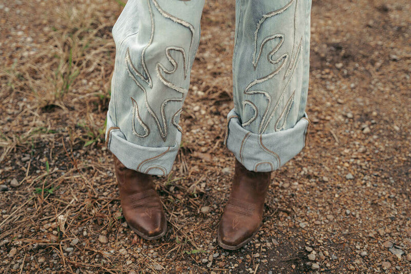 A web designer wearing light blue jeans with an abstract stitched pattern, jeans cuffed at the bottom, and brown leather boots stands on gravel and dirt ground—showcasing creative style worthy of Sweet Pea Ink Creative’s brand strategy.