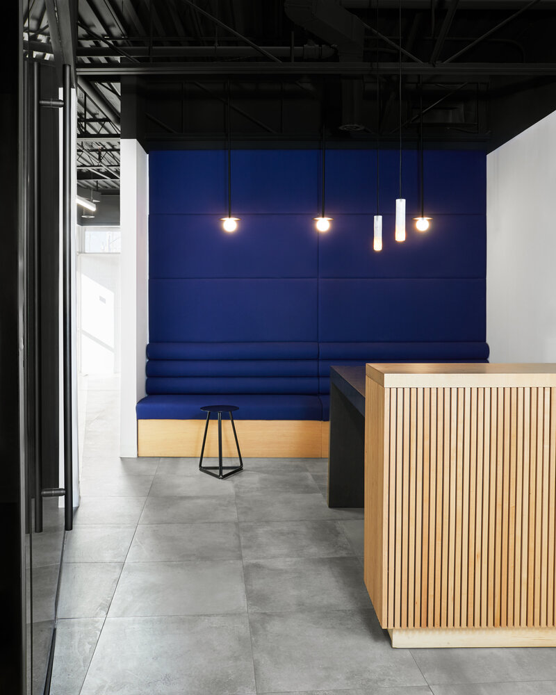 A work area with a high black ceiling, grey tiled flooring, a long blue booth-style bench, a black stool and desk, and a tall paneled wood desk