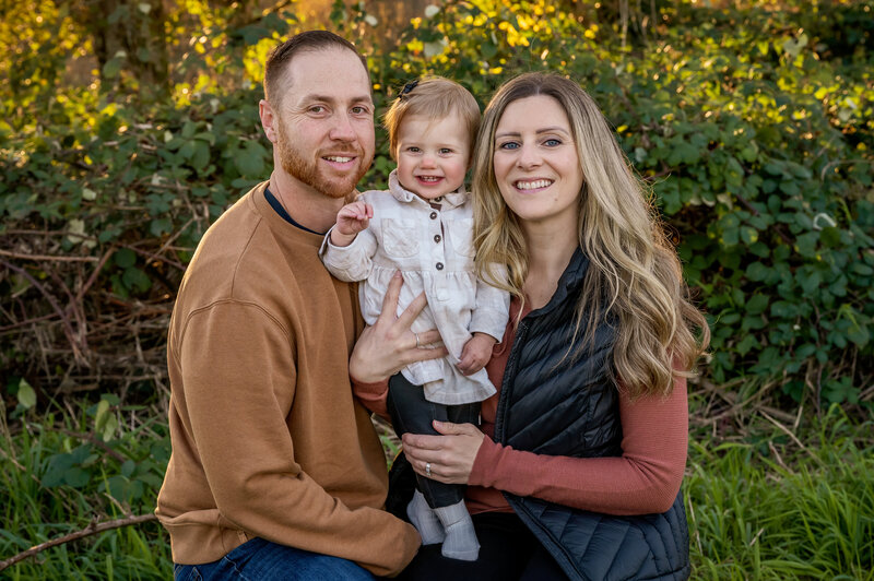 Mom and dad with their toddler smiling in a field of greenery at Pitt Lake in the fall.