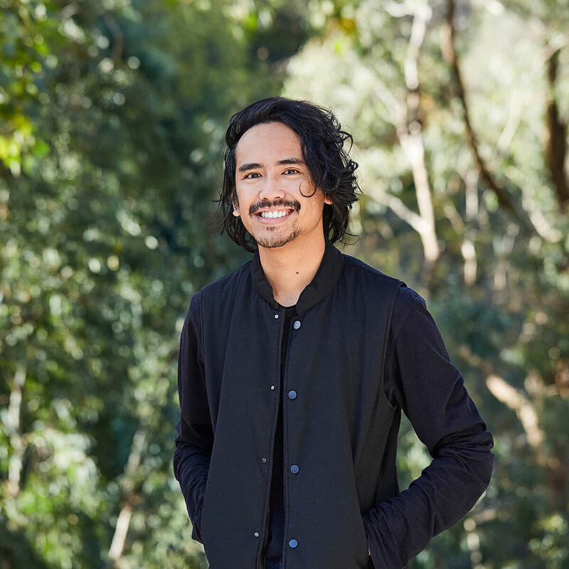 Smiling man outside with hands in pockets.  Background is sunny Australian bush.