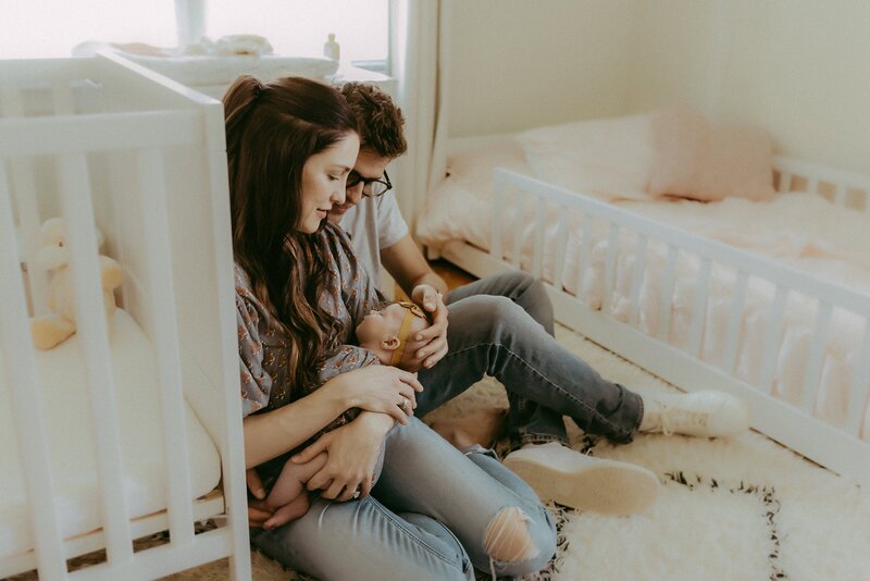 A person in a black sweater and jeans sits backward on a white chair, gazing at the camera with the distinct style of a  Macon newborn photographer, set against a plain backdrop.