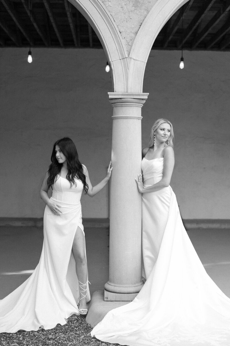 Two brides in satin gowns posing on either side of a stone column.
