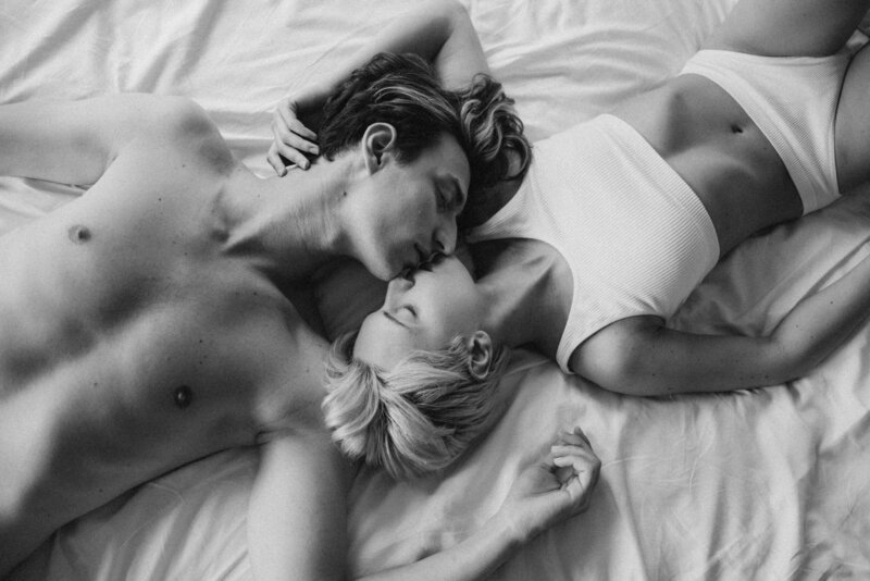 black and white image of man and woman kissing while laying on bed sheets