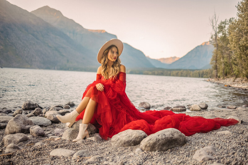 woman in a long red dress sitting on a rock by the lake with mountains in the background