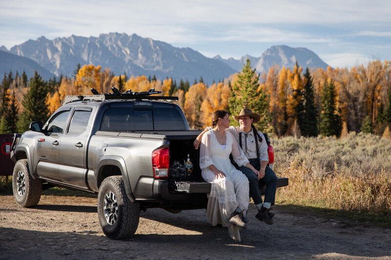 Two brides sit on the tailgate of a truck, surrounded by golden aspens and mountain views in autumn