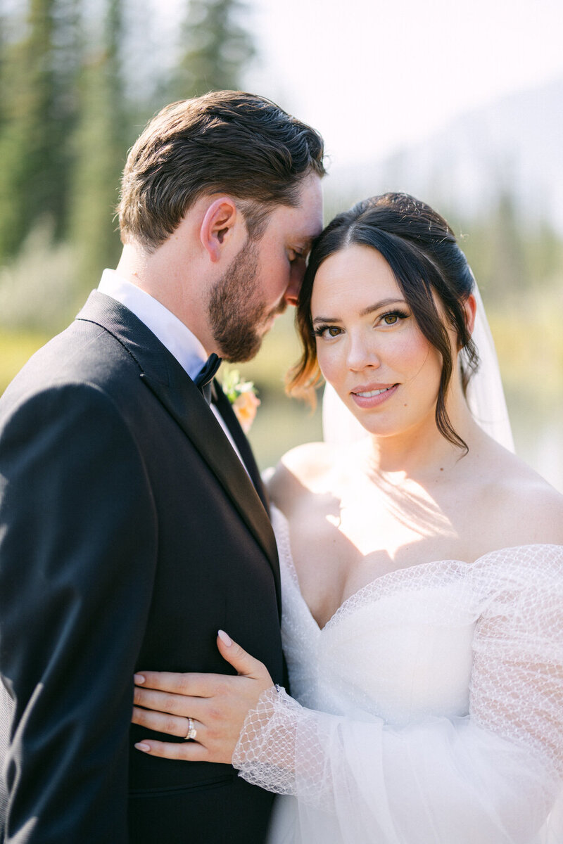 This elegant photograph captures the bride looking directly into the camera while the groom stands beside her by the Bow River in Canmore, Alberta. The stunning natural backdrop and the bride’s captivating gaze create a timeless and beautiful image of their special day.