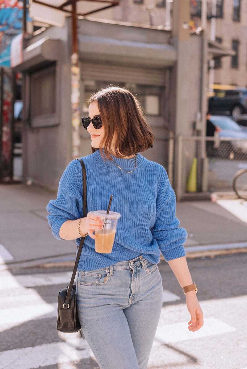 Madison walks with iced coffee in NYC wearing a blue sweater and sunglasses—reflecting her approachable and empowering presence as a digital partner.