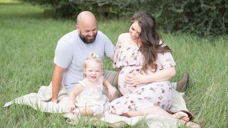 pregnant mother sitting in field smiling down to older daughter who is sitting in dad's lap